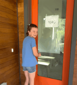 Harper stands in front of an orange door, smiling as she looks over her shoulder at the camera.